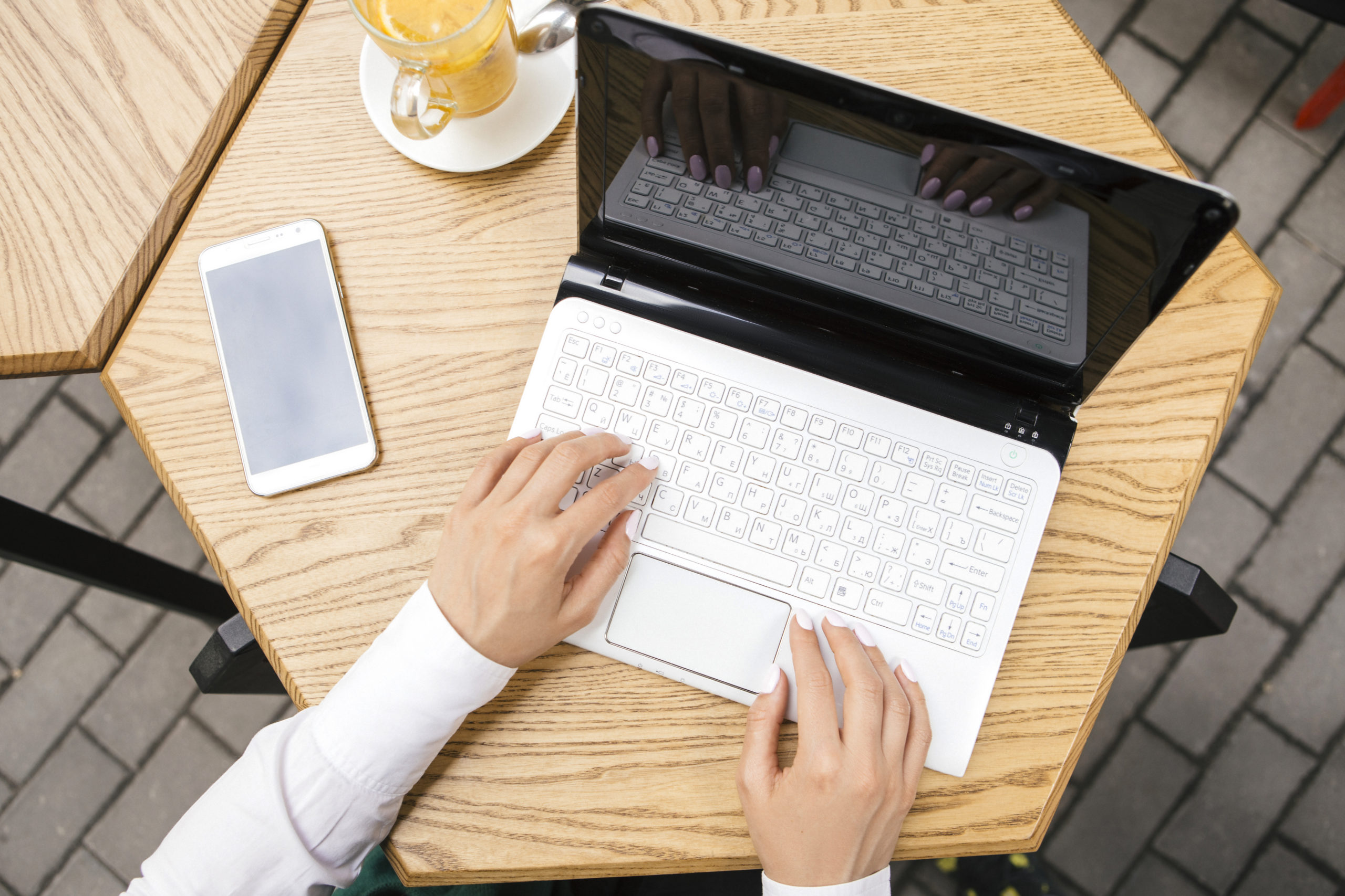 Eine Person tippt an einem weißen Laptop an einem Holztisch im Freien und recherchiert im Kosmetikstudio Hamburg, ein Smartphone und ein Glas Orangensaft stehen daneben. - KOSMETIK ATELIER Hamburg Winterhude