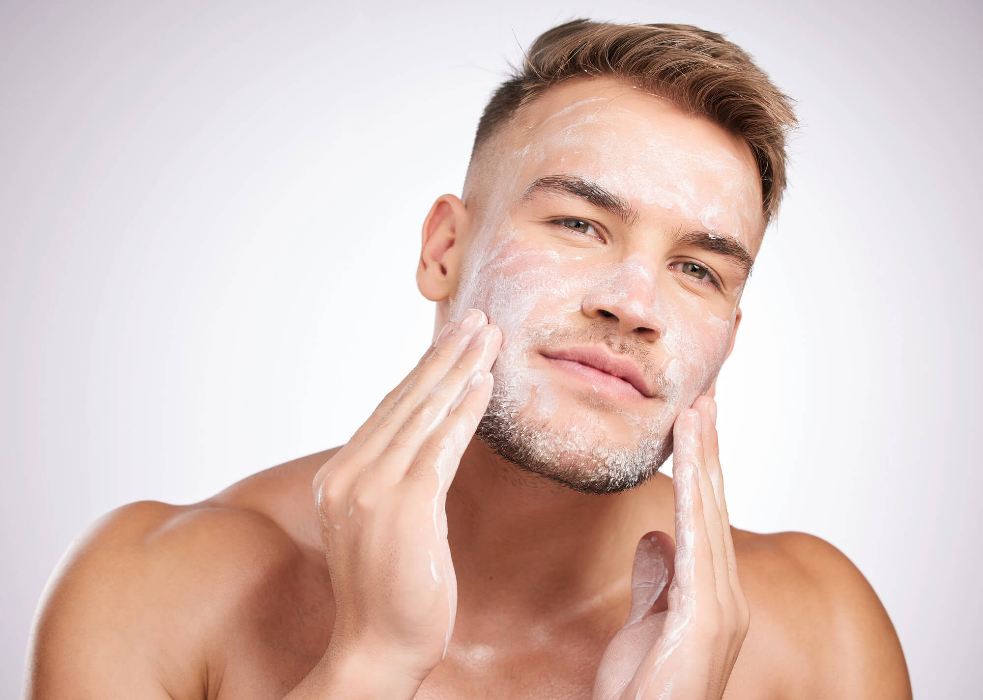 Ein junger Mann mit kurzen Haaren trägt mit beiden Händen ein weißes Gesichtsreinigungsmittel oder eine Creme auf sein Gesicht auf und blickt dabei direkt in die Kamera vor einem hellen Hintergrund. - KOSMETIK ATELIER Hamburg Winterhude